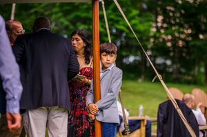 A young boy in a gray blazer stands near a wooden pole at an outdoor wedding ceremony.