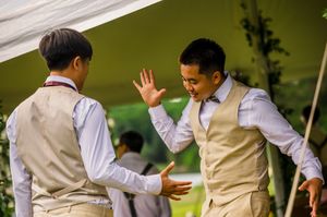 Two groomsmen in tan vests and dress shirts share a high-five at an outdoor wedding reception.