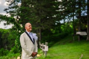 Groom in white suit smiling at outdoor wedding ceremony with floral arch and lush green trees in background.