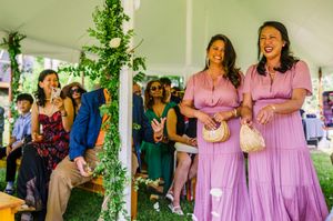 Two bridesmaids in lavender dresses smile joyfully at an outdoor wedding ceremony under a floral arch.