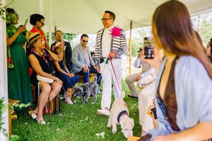 Man in striped jacket walks white dog down aisle at outdoor event while guests watch and photograph.