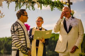 Two grooms and officiant share emotional wedding ceremony moment outdoors under floral arch.