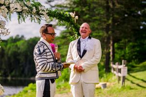 Two grooms laughing joyfully during outdoor wedding ceremony under floral arch near lake.