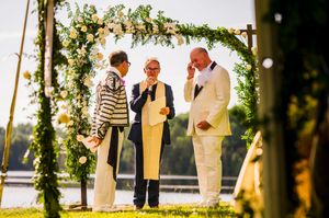 Two grooms exchange vows outdoors under a floral arch with an officiant, lakeside wedding ceremony.
