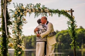 Couple sharing first kiss under floral arch with white roses and greenery at outdoor lakeside wedding ceremony.