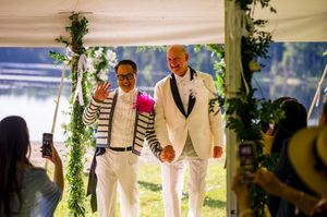 Two grooms in white suits walk hand in hand at an outdoor wedding ceremony by a lake.
