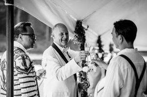 Three men in formal attire toasting with champagne glasses at an outdoor wedding reception in black and white.