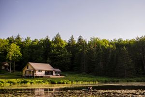 A rustic cabin sits beside a tranquil lily pad pond surrounded by lush green forest trees at golden hour.