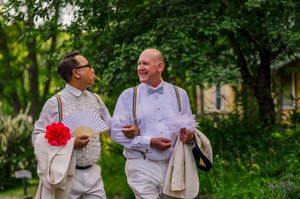 Two men in elegant white outfits with bow ties and suspenders laugh together outdoors at a garden celebration.