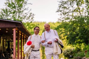 Two men in white suits walk outdoors at a wedding venue surrounded by lush green trees.