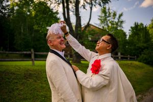 Two grooms in white suits celebrate outdoors, one adjusting the others boutonniere near lush green trees.