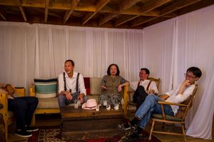Four people sitting and laughing together on a couch in a cozy, curtained room with warm lighting.