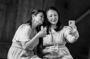 Two women in dresses take a selfie while holding drinks, smiling together in a black and white photo.
