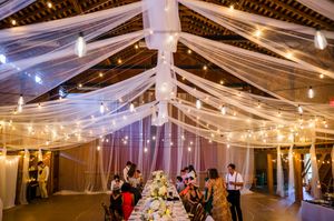 Elegant barn wedding reception with draped white fabric ceiling, string lights, and guests seated at a long decorated dining table.