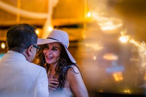 A joyful woman in a white wide-brim hat laughs with a man at a warmly lit evening celebration.