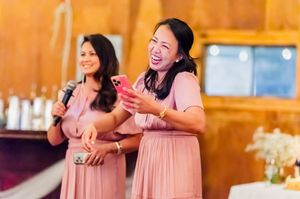 Two bridesmaids in pink dresses laughing and giving a speech at a wedding reception in a rustic venue.