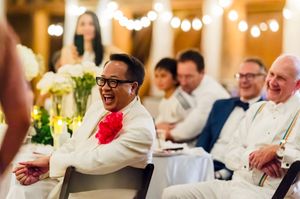 Guests laughing joyfully at a wedding reception, man in white suit with red flower boutonniere enjoying the celebration.