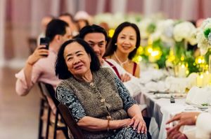 Elderly woman smiling at a wedding reception, surrounded by guests at an elegantly decorated table with white floral arrangements.