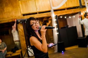 Woman singing karaoke with microphone and phone in rustic barn venue during lively party celebration.