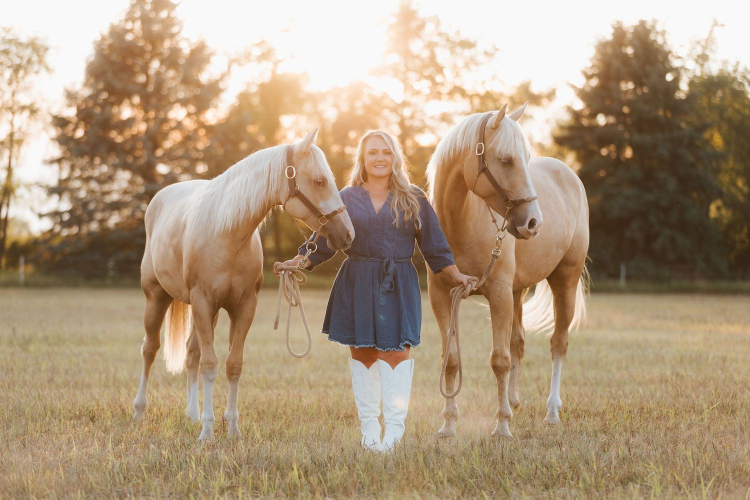 Woman in blue dress and white boots stands between two palomino horses in a sunlit field at golden hour.
