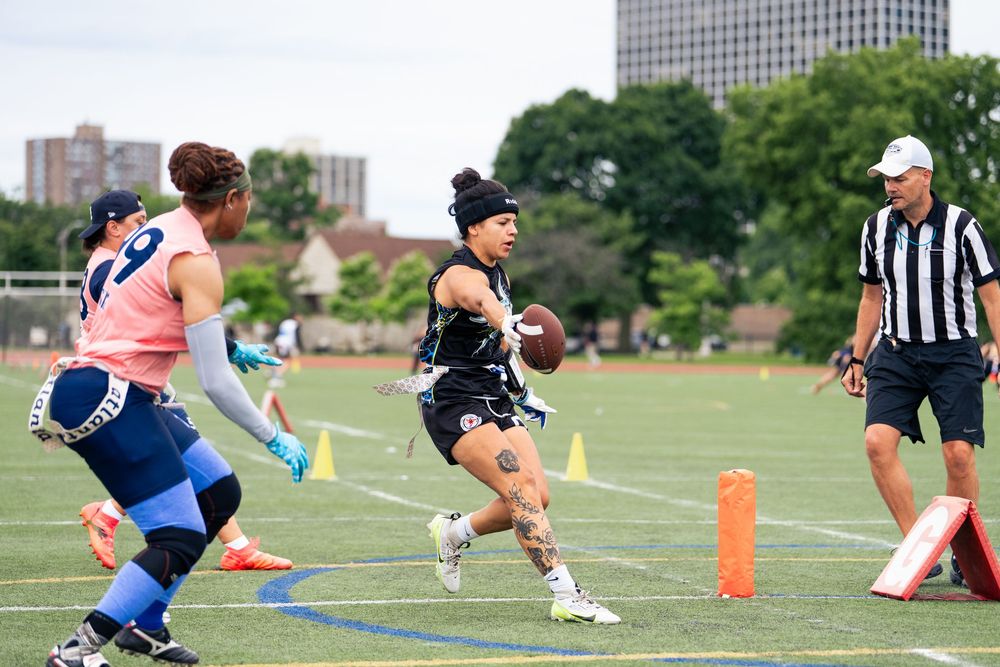 A Chicago flag football athlete completes a touchdown with a referee and opponents on defense watching at Pride Bowl hosted by NGFFL