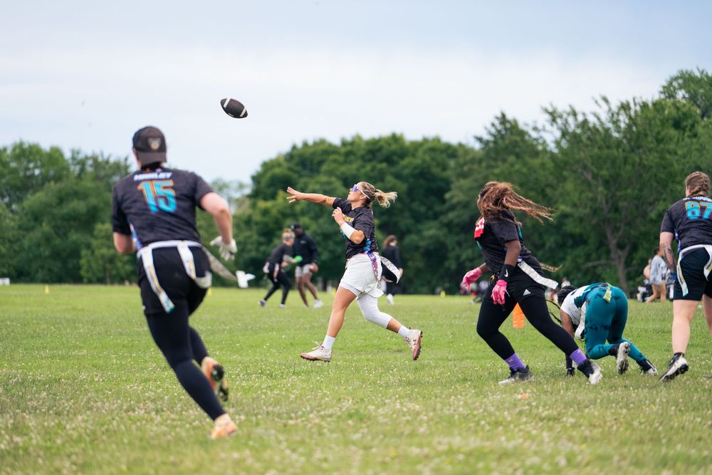 A women's+ quarterback throws the ball to make a pass on the field with an opposing rusher stumbling at Pride Bowl hosted by NGFFL