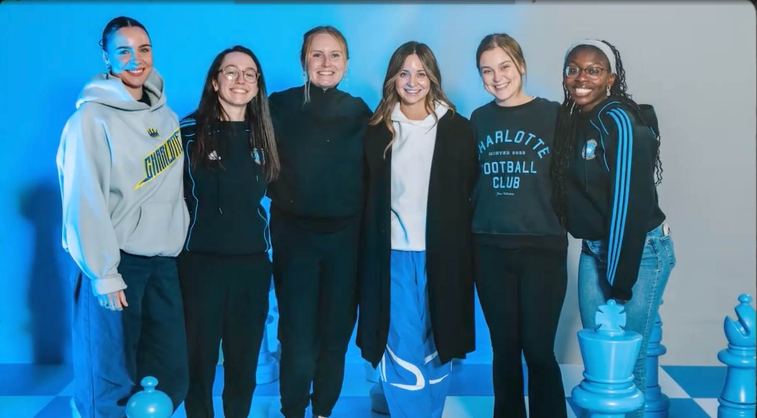 Six women pose together in front of a blue backdrop, some wearing Charlotte Football Club gear, with chess pieces visible.