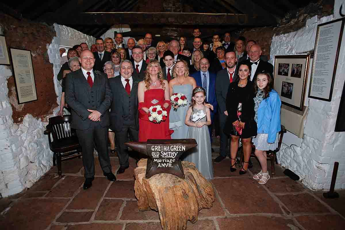 Large wedding group photo inside a rustic stone venue with bride holding red bouquet surrounded by family and guests.