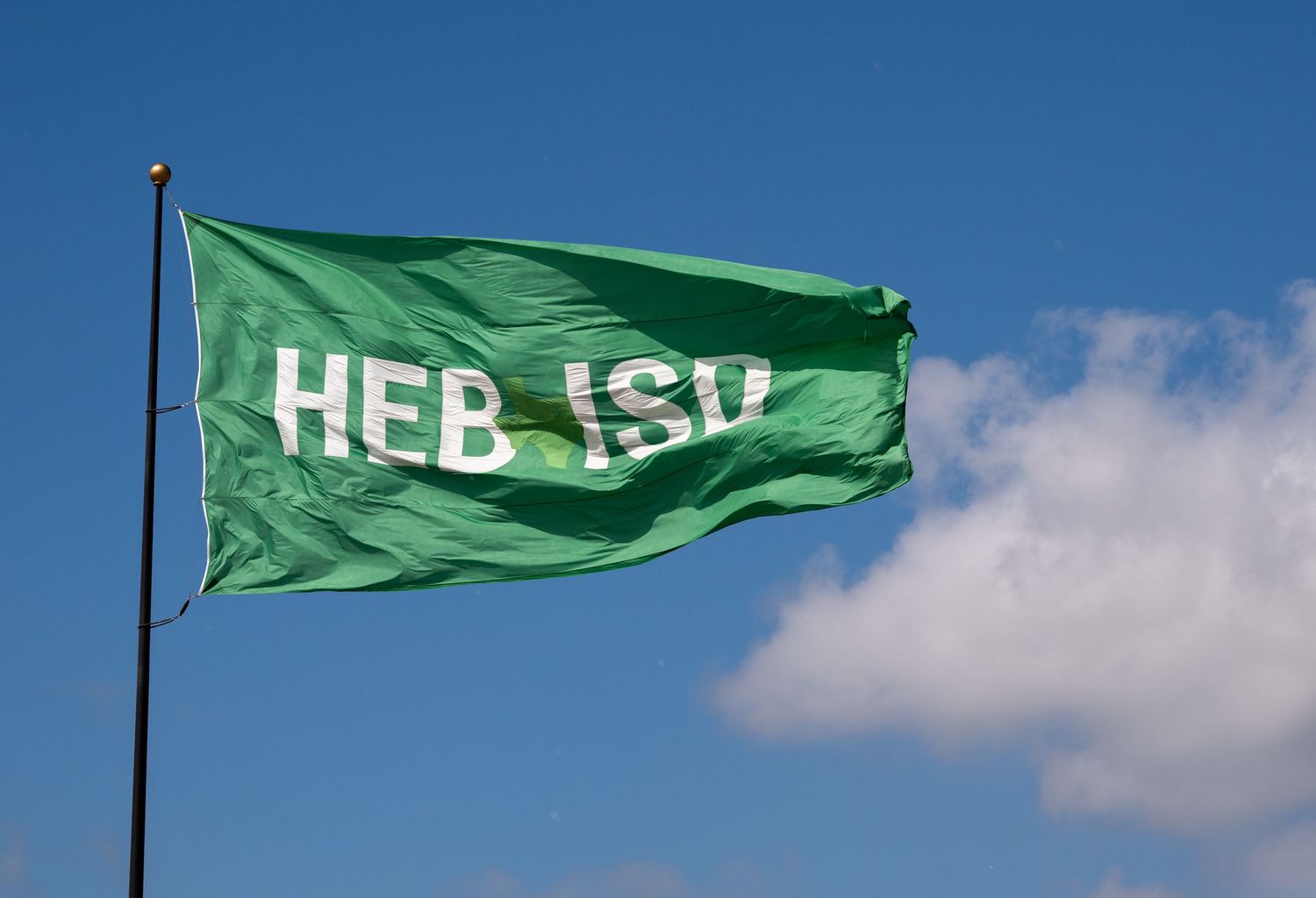 Green HEB ISD flag waving against a bright blue sky with white clouds in the background.