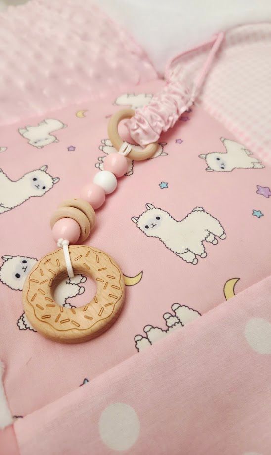 Pink baby blanket with llama pattern beside a wooden donut teether toy with beads on a string.