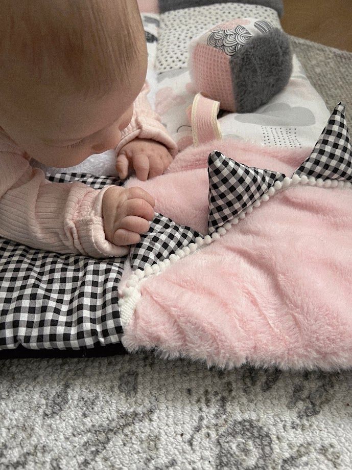 Baby girl in pink outfit exploring a soft pink and black gingham patchwork blanket with pompom trim on a gray rug.