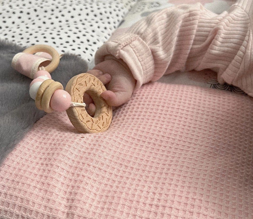 Baby hand holding a wooden donut teether rattle on a pink waffle blanket.