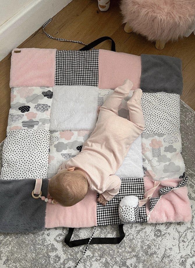Baby lying on a pink, gray, and white patchwork play mat with cloud patterns on a carpeted floor.