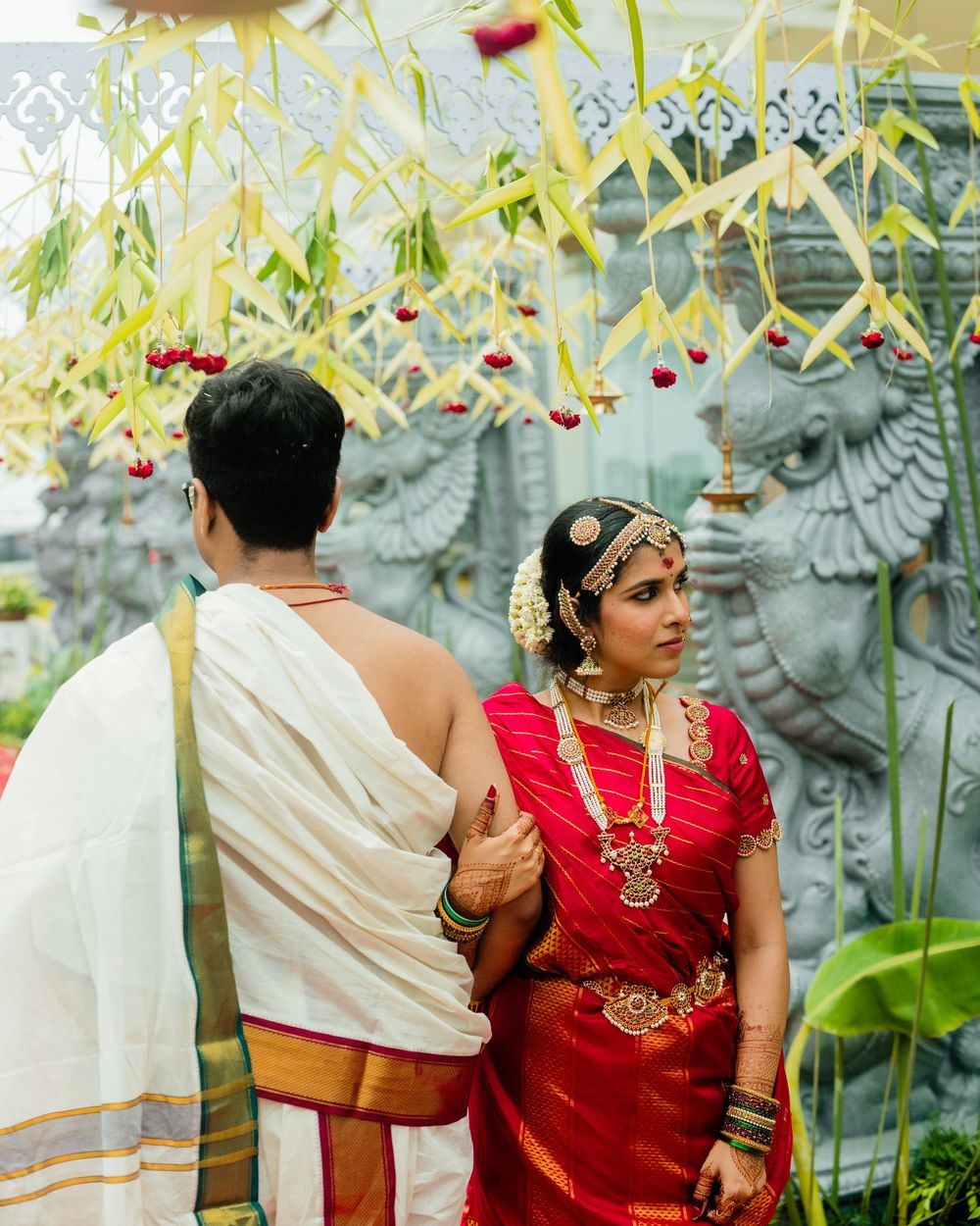South Indian bride in red silk saree with gold jewelry stands with groom in white dhoti at traditional Hindu wedding.
