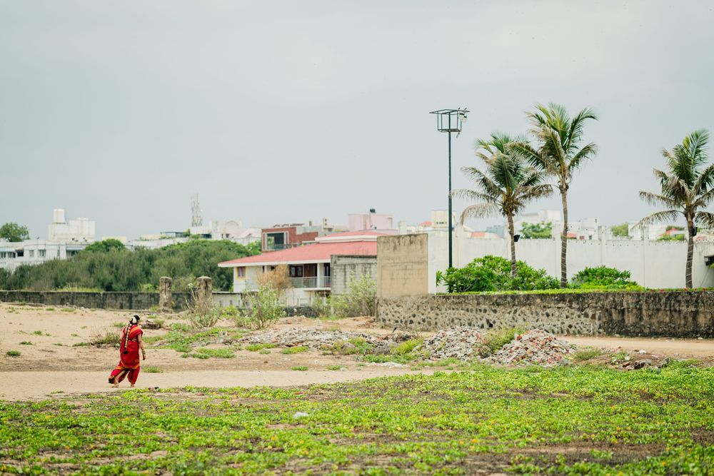 Person in red walking on sandy terrain near coastal buildings and palm trees under overcast sky.
