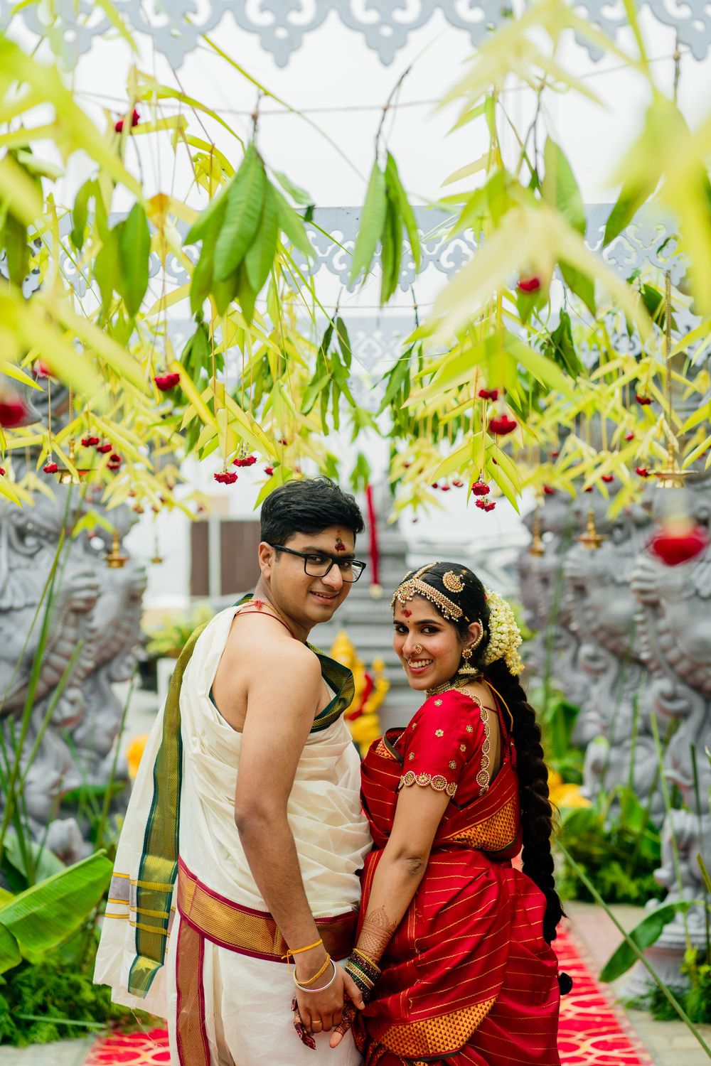 South Indian couple in traditional wedding attire holding hands under mango leaf decorations at ornate temple.