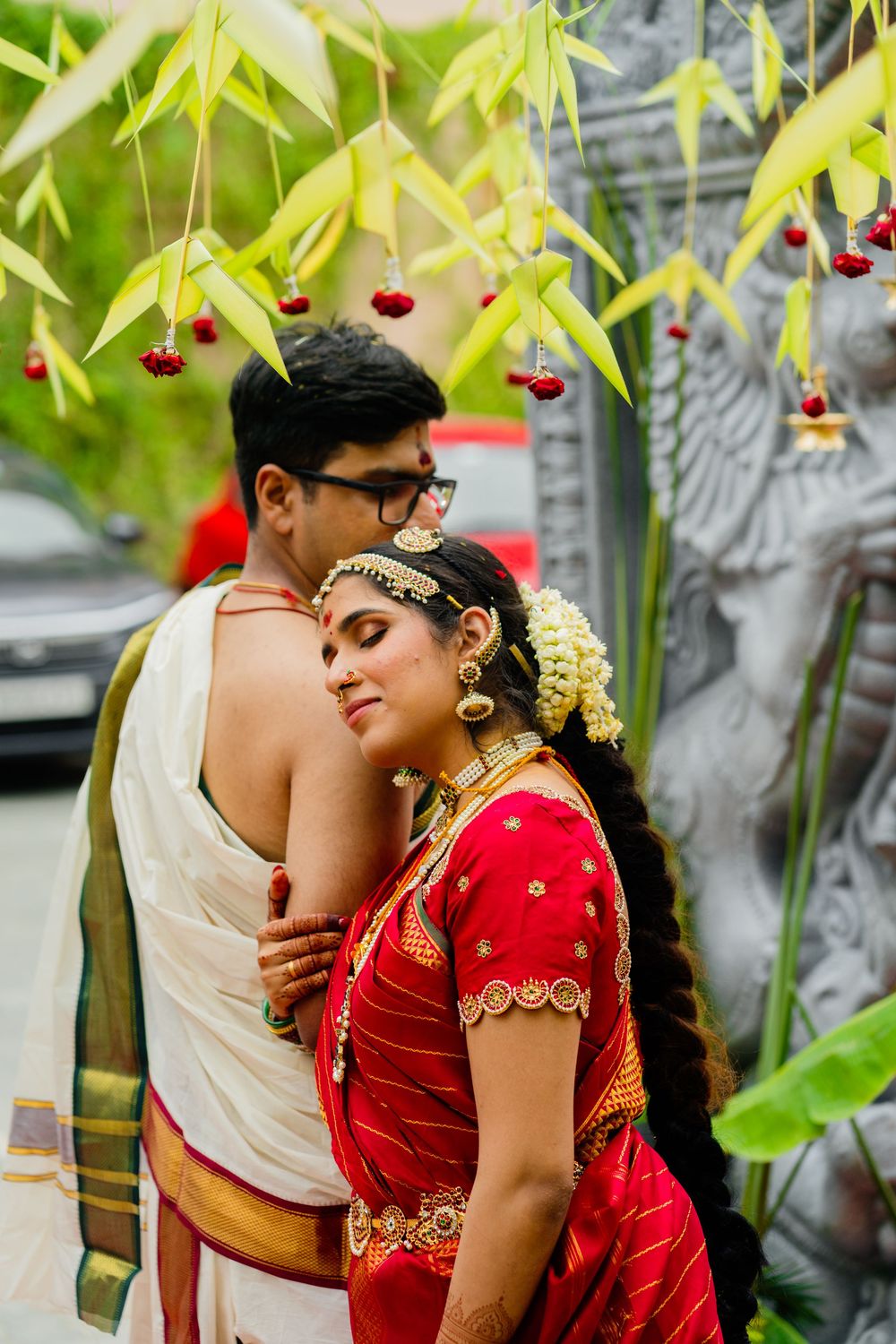 Indian couple in traditional wedding attire embracing under decorated mango leaves with red flowers.