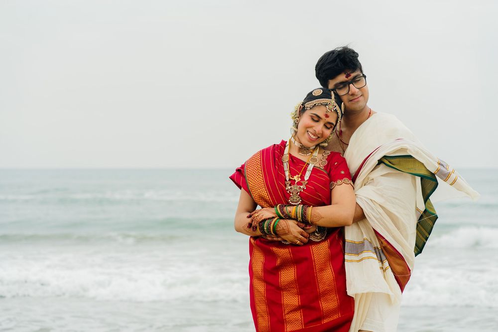 Couple in traditional Indian attire embracing on a beach, woman in red saree with gold jewelry, man in white dhoti.