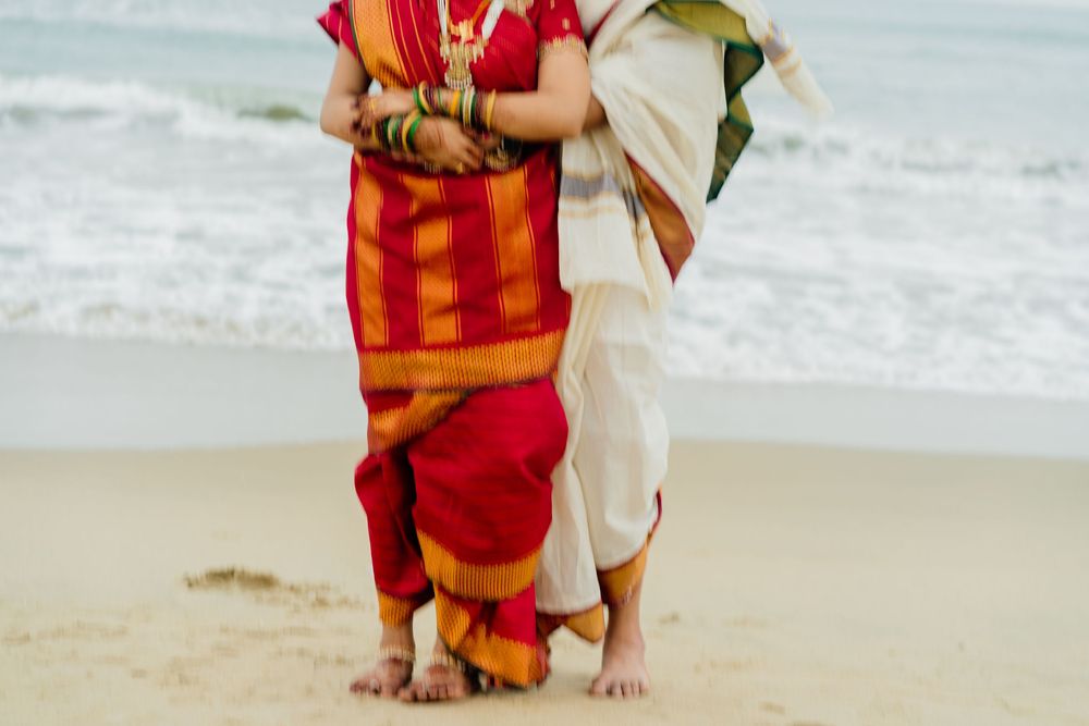 Two women in vibrant traditional Indian sarees standing barefoot on a sandy beach with ocean waves in the background.