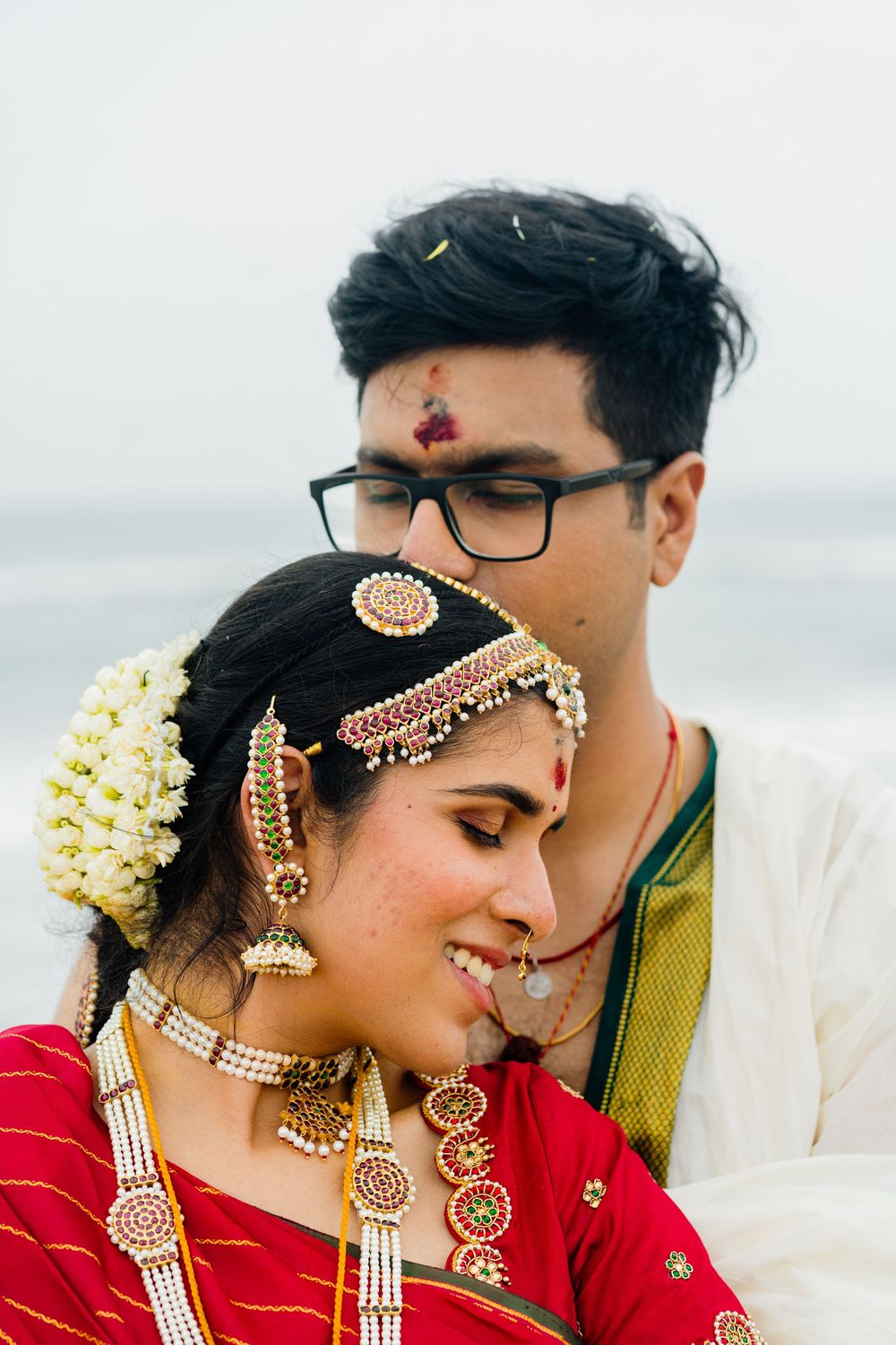 Indian bride in red saree with gold jewelry and groom in white kurta pose together at a beach wedding.
