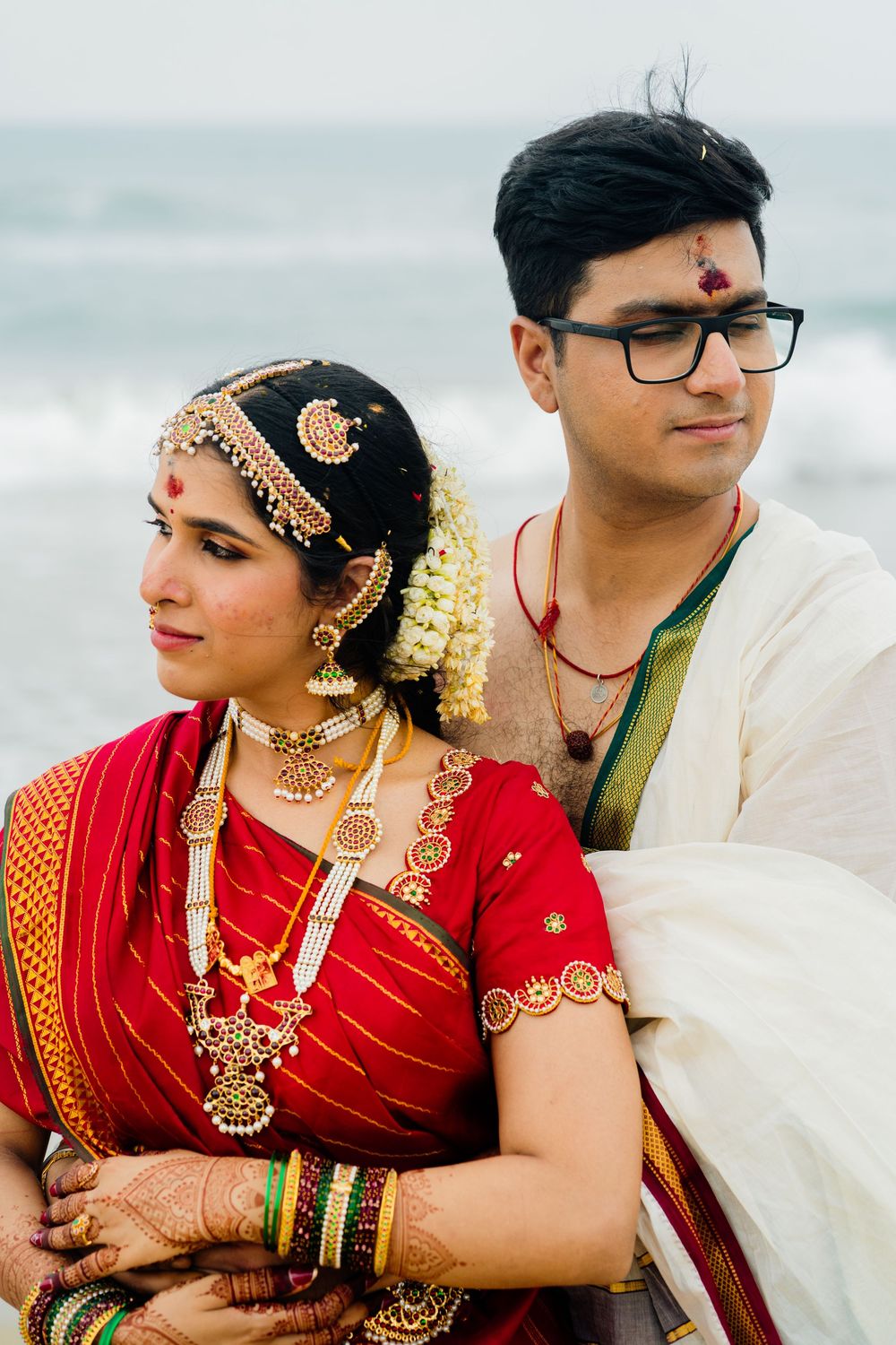 South Indian bride in red saree with gold jewelry embraces groom in white dhoti at beach wedding.