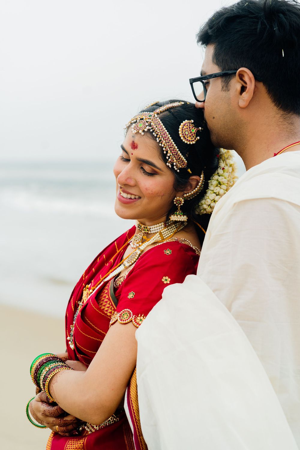 Smiling South Indian bride in red saree with floral hair accessories shares romantic moment with groom at beach.