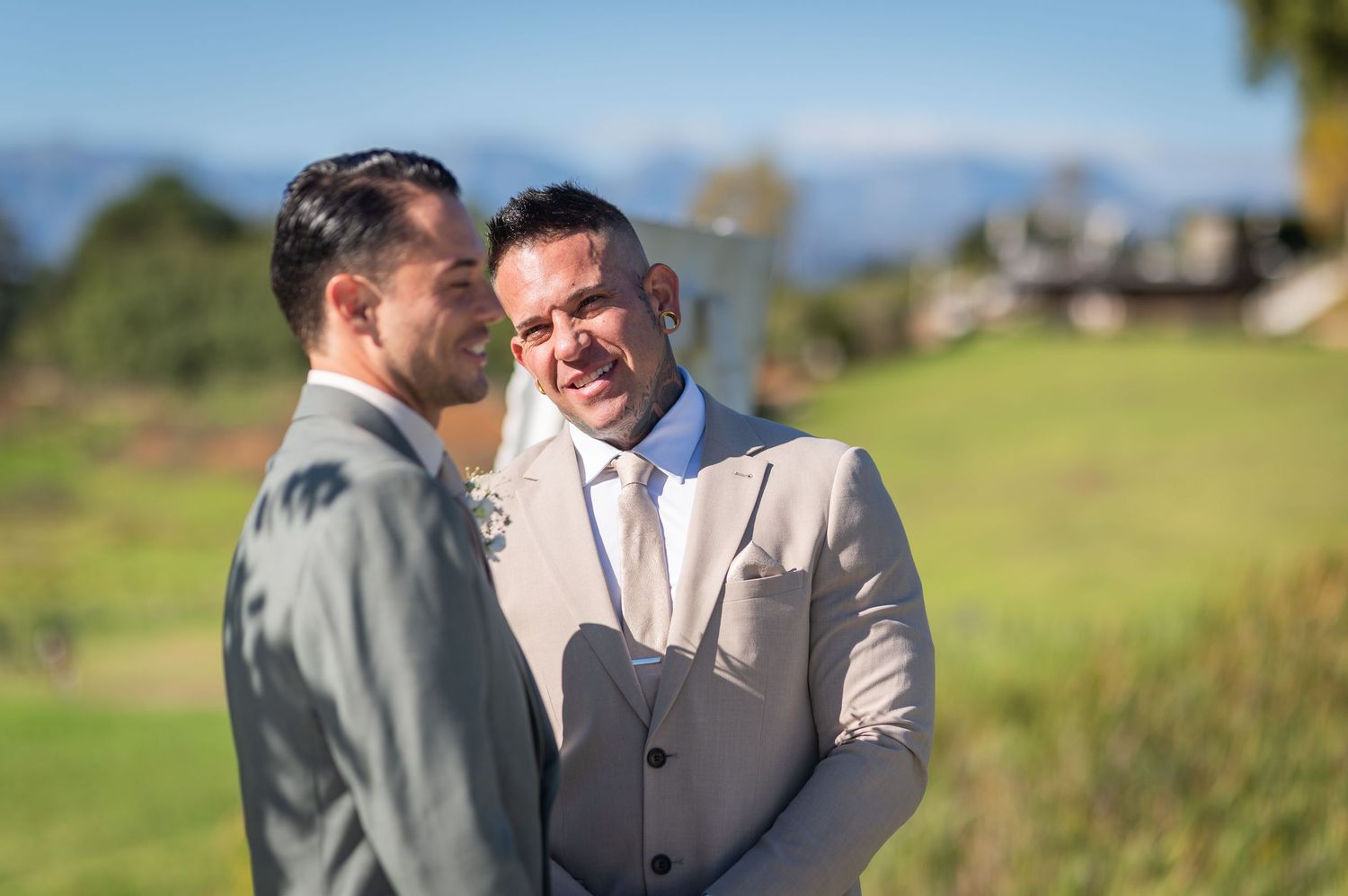 Two grooms in suits smiling at each other outdoors on their wedding day with a scenic green landscape behind them.