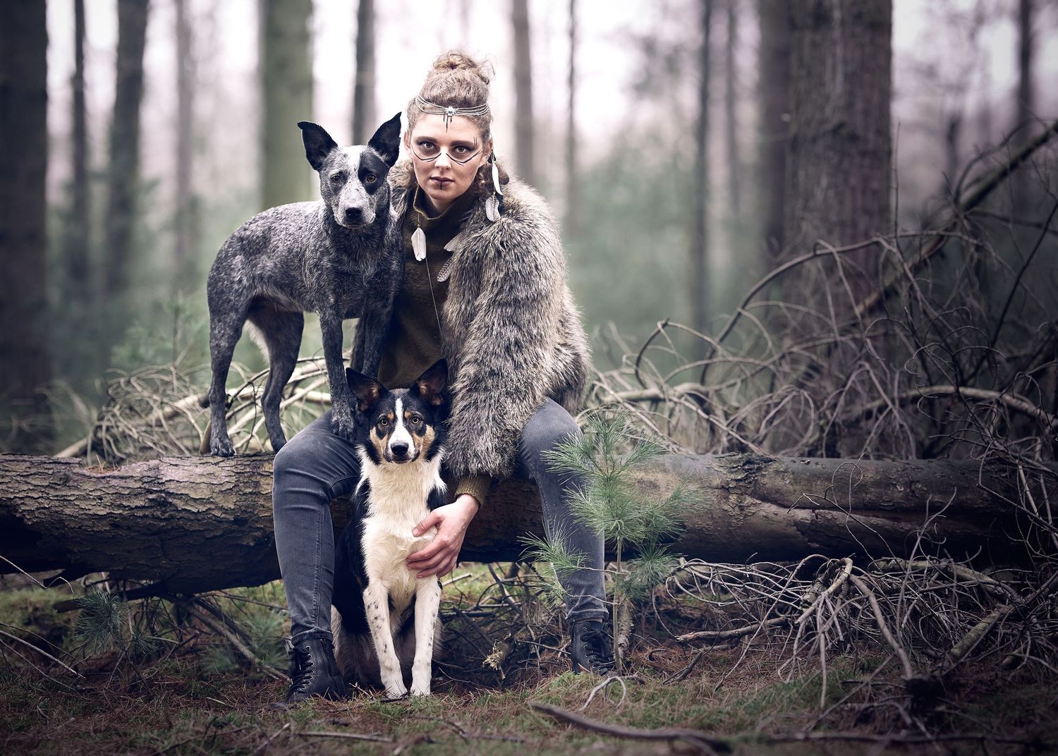 Woman in fur vest poses with two dogs on fallen log in misty forest setting.