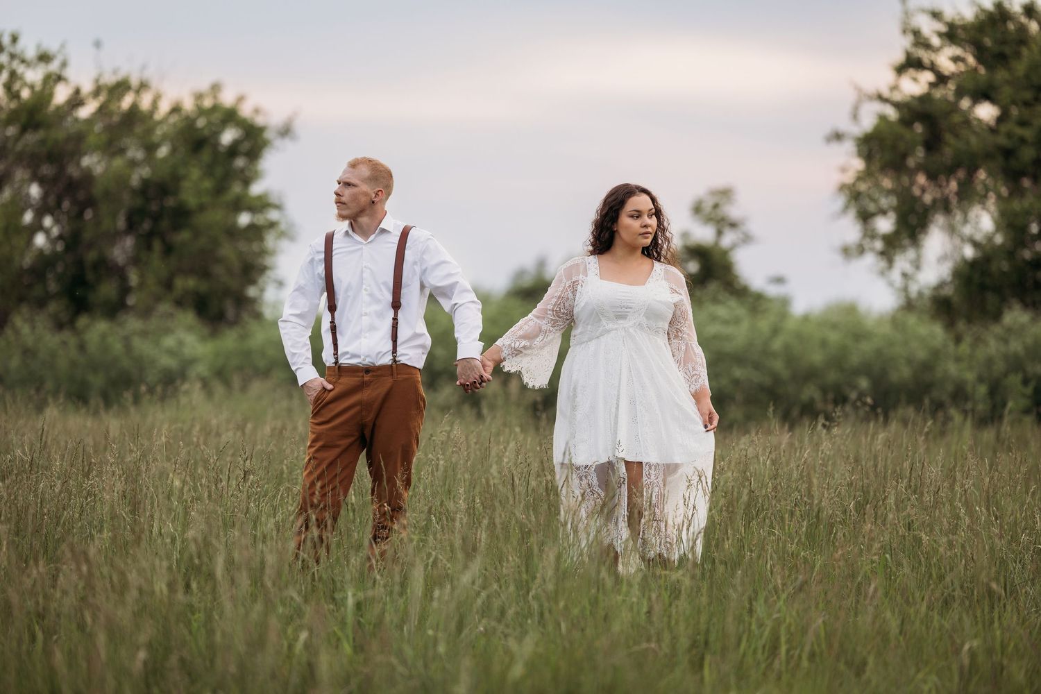 Couple sharing a romantic kiss in a lush green field at sunset with a dramatic cloudy sky backdrop.