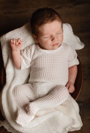 Sleeping newborn baby in white knit outfit resting peacefully in a wooden bowl with a soft white blanket.