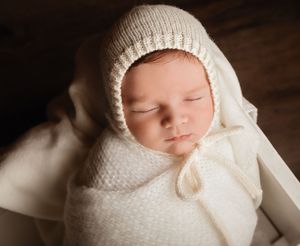 Newborn baby sleeping peacefully wrapped in cream knit blanket and bonnet in a white wooden box.