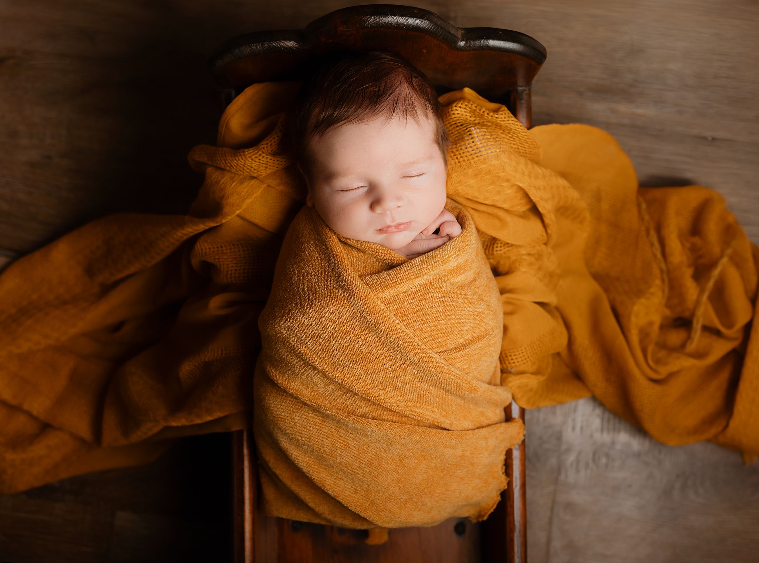 Newborn baby swaddled in a golden mustard wrap, sleeping peacefully in a small wooden bed with matching fabric.