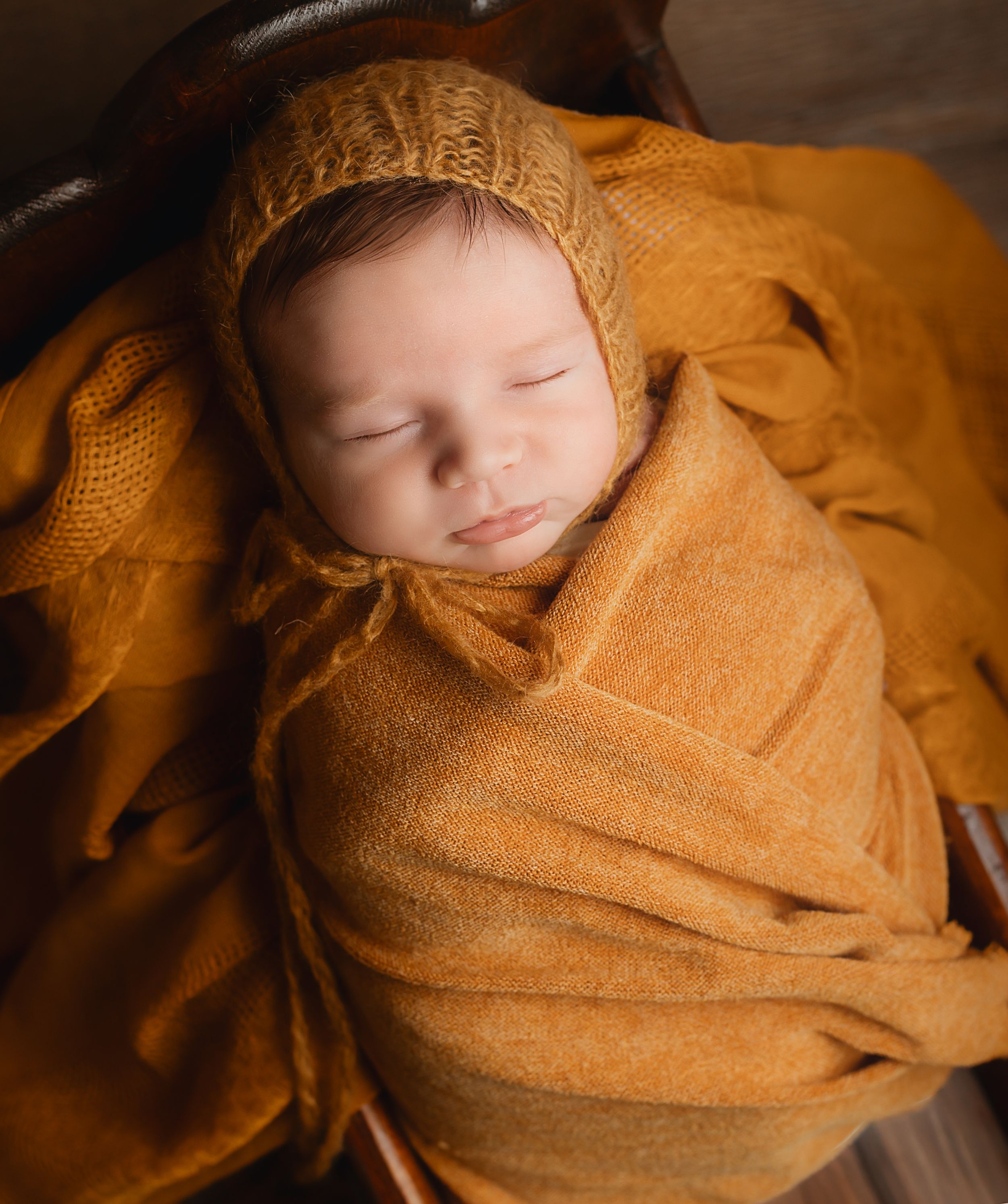 Sleeping newborn baby wrapped in a mustard yellow knit blanket and bonnet, nestled in a basket.