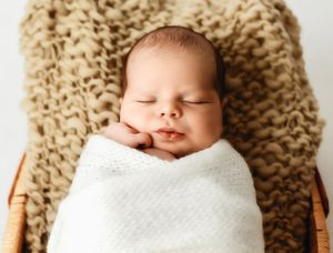 Peaceful newborn baby swaddled in white wrap sleeping in wicker basket with cozy knit blanket backdrop.